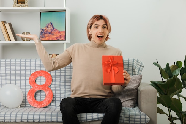 Handsome guy on happy women day holding present sitting on sofa in living room