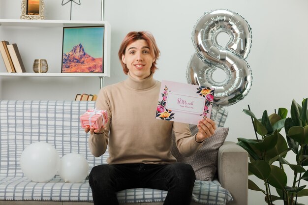 Handsome guy on happy women day holding present with postcard sitting on sofa in living room