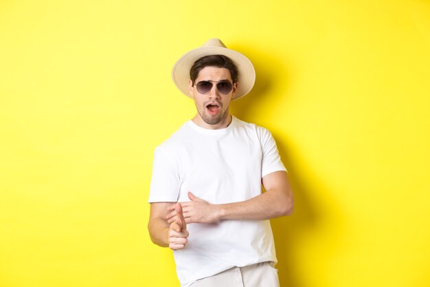 Handsome guy making finger gun shot and looking sassy, dressed for summer vacation in straw hat and sunglasses, standing against yellow background