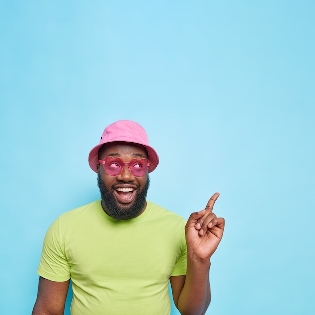 Handsome happy man with thick beard points at upper right corner dressed in summer clothes trendy pink sunglasses shows copy space for your advertisement isolated on blue wall