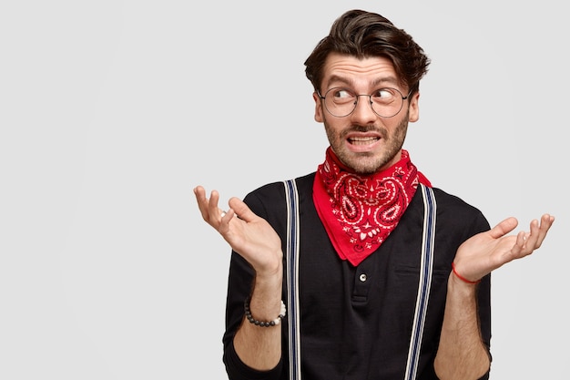 Handsome hesitant guy shrugs shoulders, looks doubtfully aside, doesn`t know what to say, wears stylish shirt and red bandana on neck