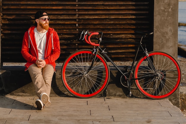 Free photo handsome hipster style bearded man in red hoodie sitting relaxing alone with backpack and bicycle in morning sunrise by the sea healthy active lifestyle traveler backpacker