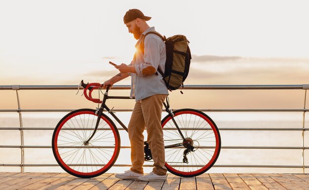 Handsome hipster style bearded man with backpack wearing denim shirt and cap traveling with bicycle in morning sunrise by the sea drinking coffee, healthy active lifestyle traveler backpacker