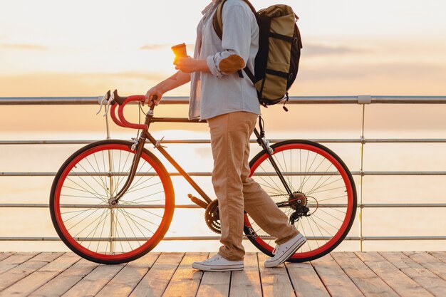 Handsome hipster style bearded man with backpack wearing denim shirt and cap with bicycle in morning sunrise by the sea drinking coffee, healthy active lifestyle traveler backpacker