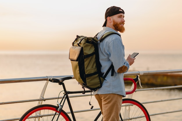 Handsome hipster style bearded man with backpack wearing denim shirt and cap with bicycle in morning sunrise by the sea drinking coffee, healthy active lifestyle traveler backpacker