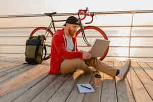 Free photo handsome hipster style bearded man working online freelancer on laptop with backpack and bicycle in morning sunrise by the sea healthy active lifestyle traveler backpacker