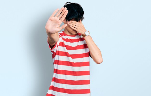 Handsome hipster young man wearing casual striped t shirt covering eyes with hands and doing stop gesture with sad and fear expression. embarrassed and negative concept.