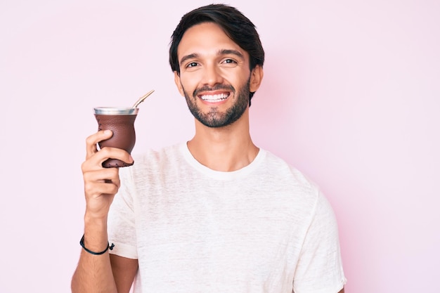 Free photo handsome hispanic man drinking mate infusion looking positive and happy standing and smiling with a confident smile showing teeth