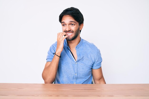 Handsome hispanic man wearing casual clothes sitting on the table looking stressed and nervous with hands on mouth biting nails anxiety problem