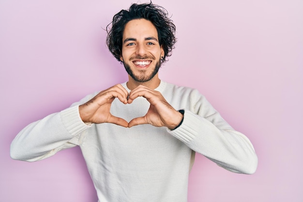 Handsome hispanic man wearing casual white sweater smiling in love doing heart symbol shape with hands. romantic concept.