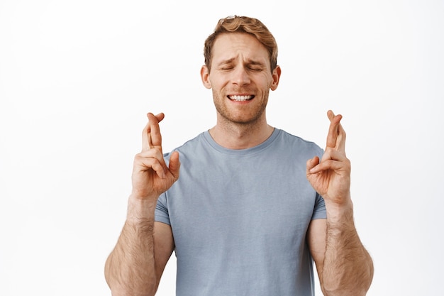 Free Photo handsome hopeful ginger guy praying, close eyes and cross fingers for good luck, making wish, waiting for sign, anticipating news, standing over white wall