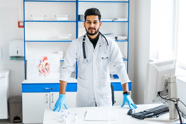 A handsome Indian doctor man in a white medical gown and with a stethoscope near his workplace