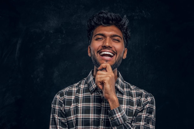 Free photo handsome indian guy in plaid shirt smiling and holds his hand on chin. studio photo against a dark textured wall