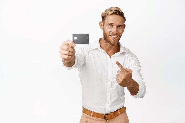 Handsome macho man pointing and showing credit card bank product advertising big sale promo offer standing over white background