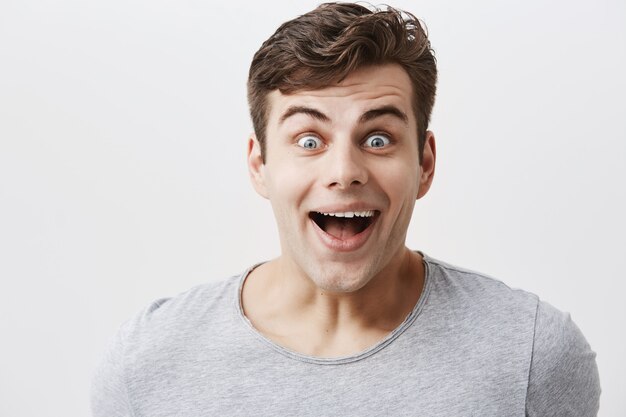 Handsome male in gray t-shirt, smiles with surprisment, looks with bugged blue eyes, being astonished to hear unexpected news or see gift from friend. Positive emotions and feelings concept.