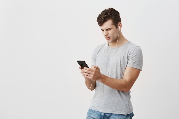 Free Photo handsome male student holds mobile phone in his hands, looks with confused expression at screen, wears white earphones, frowns face. caucasian man confused with the message he got from girlfriend.