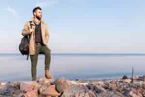 Free photo handsome male traveler standing in front of sea holding handbag on shoulder looking away