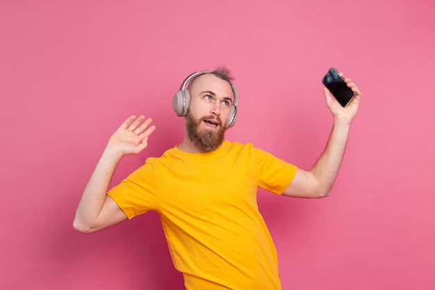 Handsome man in casual dancing with mobile phone and headphones isolated on pink background