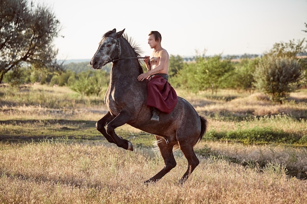 Handsome man cowboy riding on a horse - background of sky and trees. A man in red wide pants without a shirt. Show