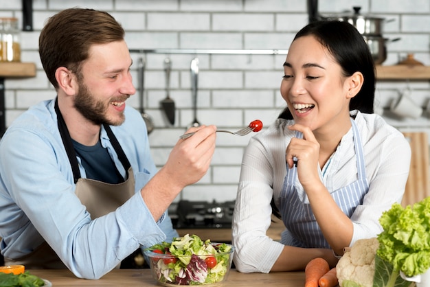 Free photo handsome man feeding cheery tomato to his wife in kitchen