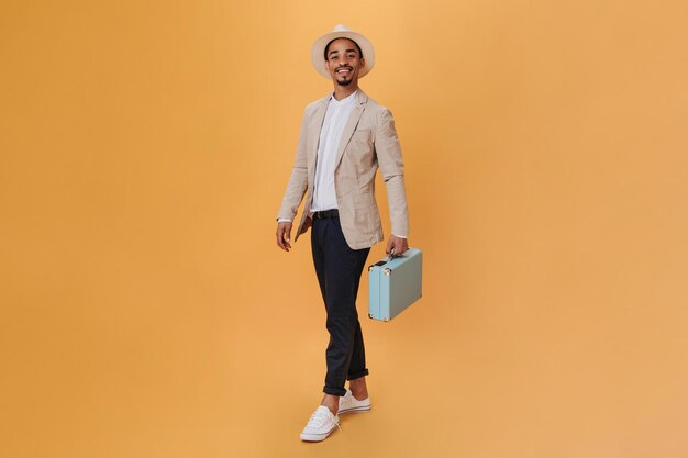 Handsome man in hat and suit posing with suitcase on orange background Young guy in beige jacket and black pants holding blue bag