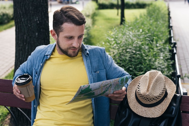 Free photo handsome man holding disposable coffee cup while reading map while sitting in park