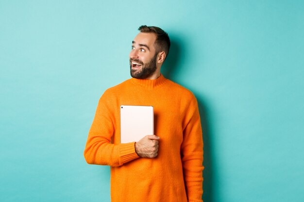 Handsome man holding laptop, looking left with surprise and amazement, standing in orange sweater