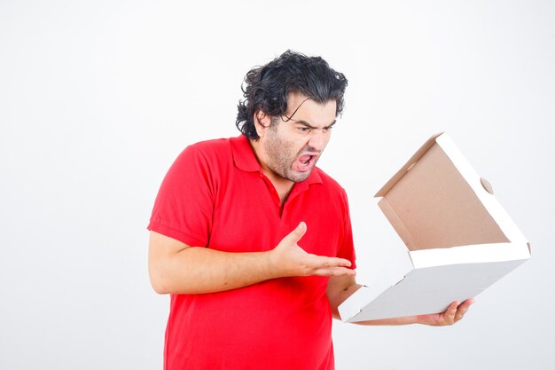 Handsome man opening paper box, stretching hand toward it with angry manner in red t-shirt and looking angry , front view.