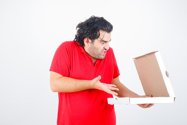 Handsome man opening paper box, stretching hand toward it with surprised manner in red t-shirt and looking shocked , front view.