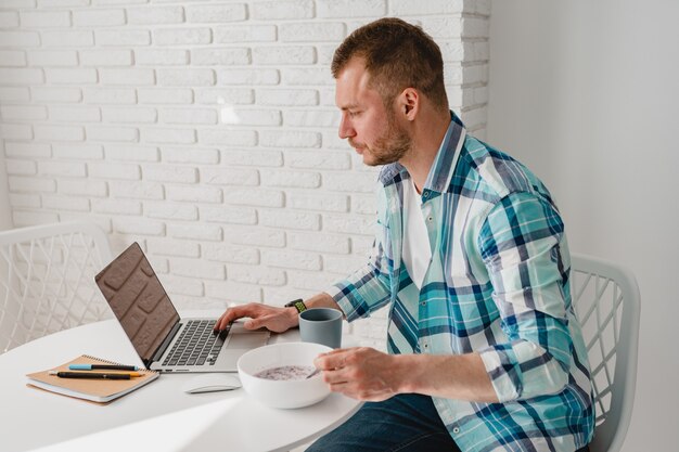 Handsome man in shirt having breakfast at home at table working online on laptop from home