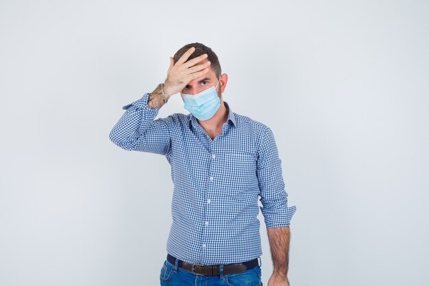 Handsome man in shirt, jeans, mask holding hand on head, having headache and looking exhausted , front view.