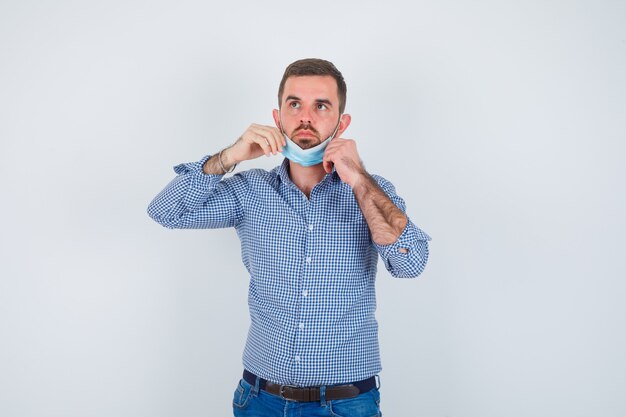 Handsome man in shirt, jeans, mask holding mask under chin and looking serious , front view.