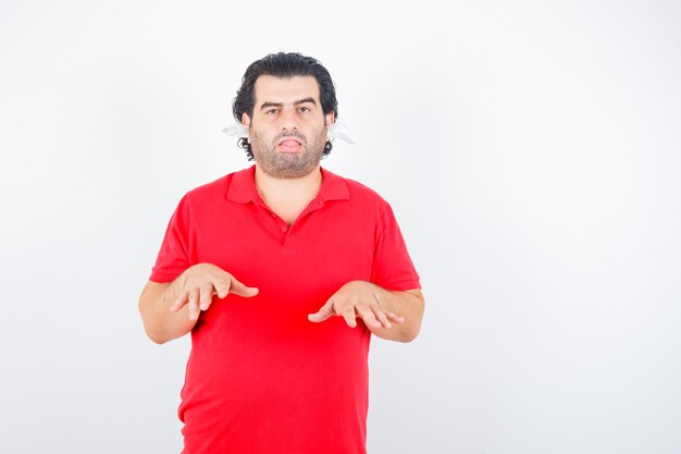 Handsome man showing height gesture, standing with napkins in ears in red t-shirt and looking tired , front view.