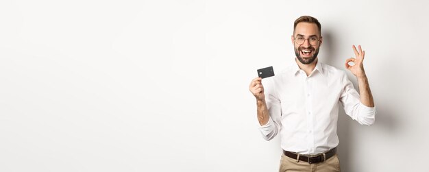 Handsome man showing his credit card and okay sign recommending bank standing over white background