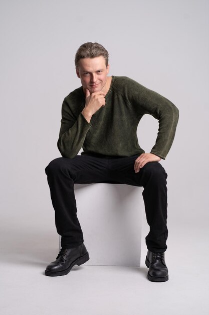 Handsome man sitting on the white cube with hand touching his chin looking at camera and smile wearing black denim and greengray knitted jumper on white background