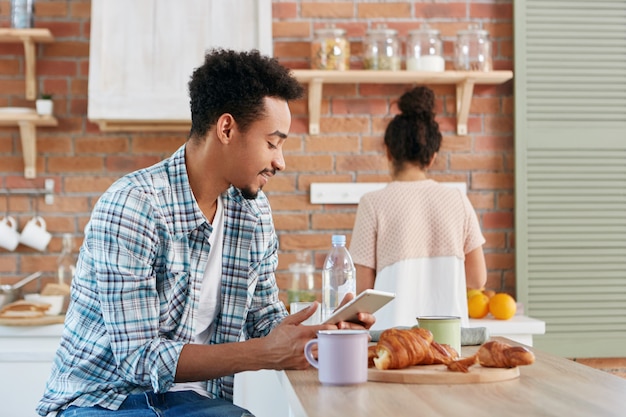 Handsome man spends morning at kitchen, has breakfast, reads electronic book or texts with friends using digital tablet