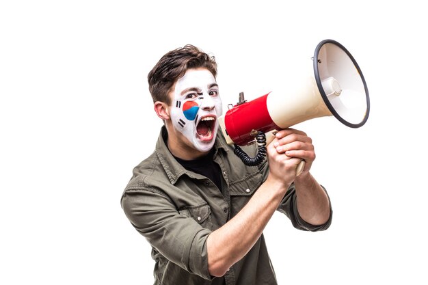 Handsome man supporter loyal fan of Korea Republic national team with painted flag face screaming into megaphone with pointed hand. Fans emotions.