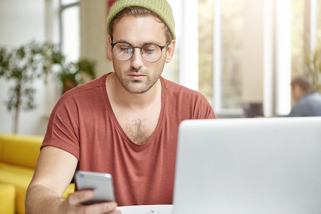 Free photo handsome man wears trendy round spectacles, hat and t shirt, textes message on smart phone