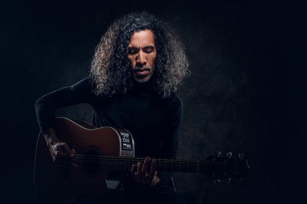 Free photo handsome man with long hair is playing acoustic guitar at dark studio.