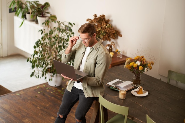 Free photo handsome manager man in glasses sits on table holds laptop and types sends message works on project