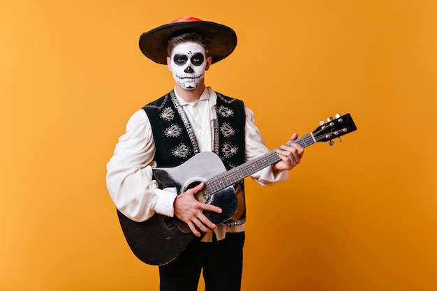 Free Photo handsome mariachi with zombie makeup standing on yellow wall. inspired man in sombrero playing guitar in halloween.