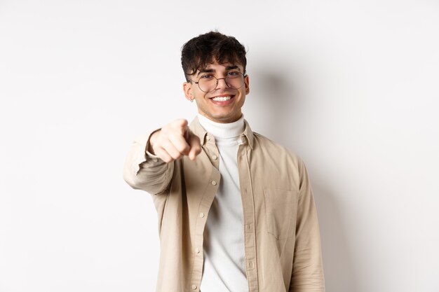 Handsome modern guy in glasses pointing at camera, smiling and choosing you, recruiting or inviting to event, standing on white background.