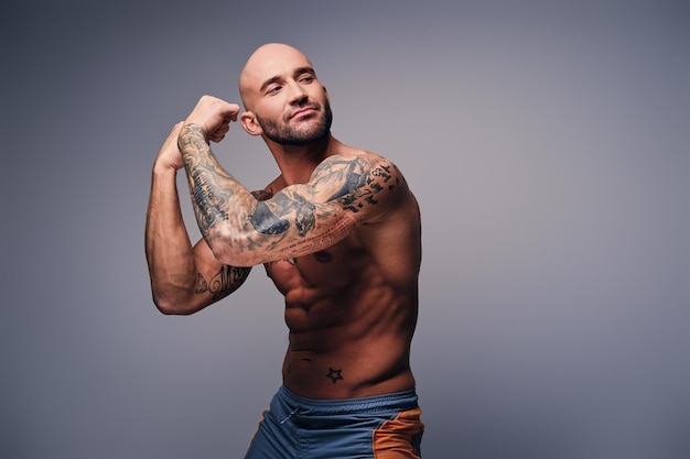 Free photo handsome muscular shaved head male with tattooed torso shows his muscles over grey background.