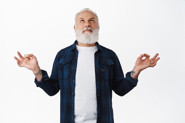 Free Photo handsome old man with tattoos meditating, smiling while looking up and making zen mudra gesture, practice yoga, breathing peaceful, relaxing mind, standing over white wall