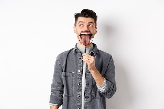 Handsome positive guy showing white perfect teeth and tongue with magnifying glass, looking left at logo, standing against white background.