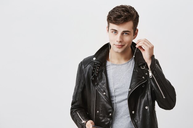 Handsome positive man in black leather jacket with faint smile, listens music in earphones. Young caucasian male enjoys pleasant melodies, wearing white earphones, looking  with blue eyes.