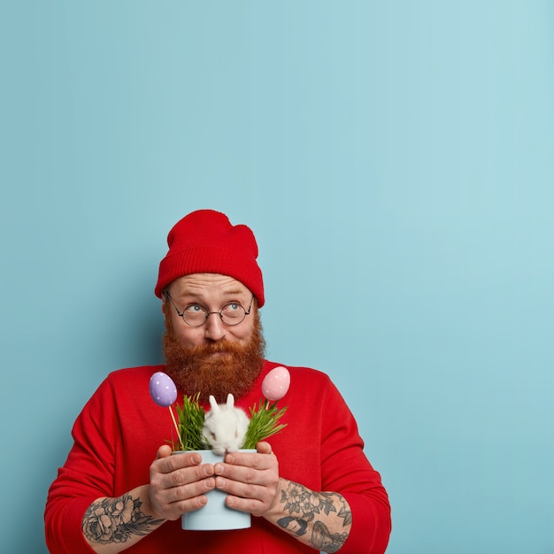 Free photo handsome positive man with thick ginger beard looks pensively above, thinks how to celebrate easter, carries traditional bunny in pot with grass and eggs, has tattoo, wears stylish red clothes