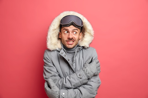 Free photo handsome puzzled unshaven man skier shievers and trembles during cold frosty day hates winter cold wears snowboard goggles and grey jacket.