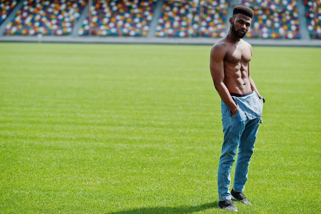 Handsome sexy african american muscular bare torso man at jeans overalls posed at green grass of stadium football field Fashionable black man portrait