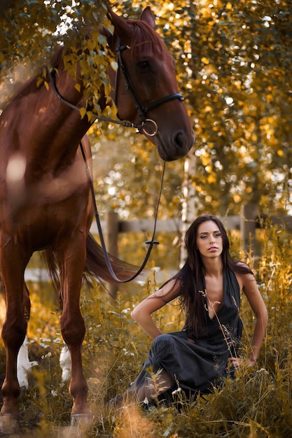 Free photo handsome slim woman sitting on the ground with brown horse near her.